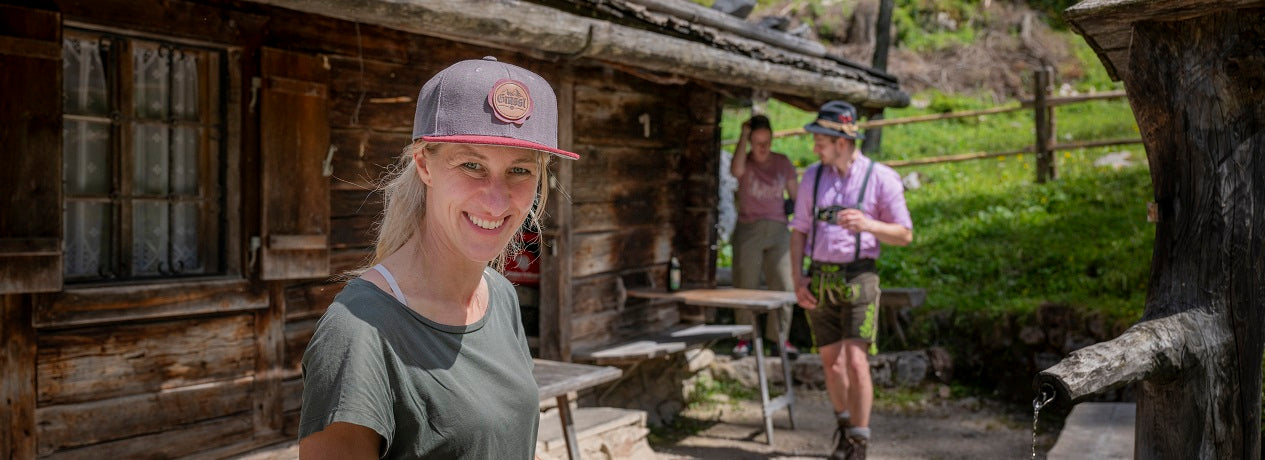 Frau mit Grassl Cap. Im Hintergrund ist eine Brennhütte mit Bergbrenner.